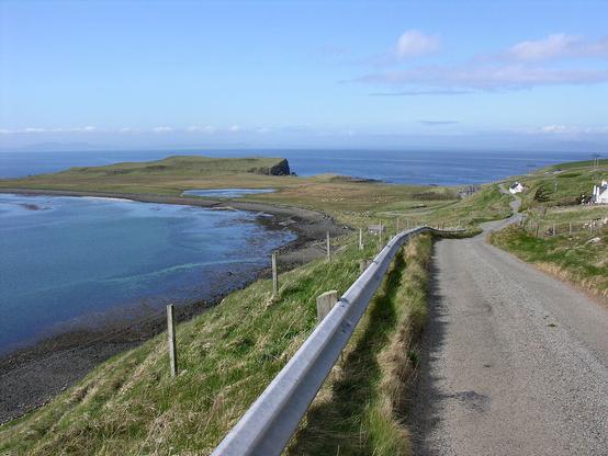 Coastal scenery at Trumpan. The image shows a narrow road leading up the right side of the frame with a metal crash barrier to its left. The ground drops steeply to the shore on the left with a calm sea and a stony beach. The shore curves round to the left, to a promontory with sea this side of it and beyond it, where it continues almost to the horizon, which has a faint outline of distant land. The scene is in sunlight.