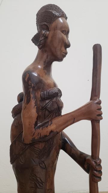 Sculpture in dark grained wood of an African woman holding a long mortar to pound maize