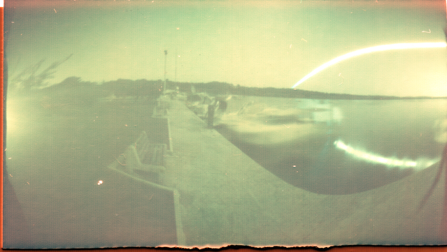 schwarzweiß Aufnahme eines bootssteg und der promenade mit ein paar booten im wasser. im hintergrund das meer und der sonnenbogen mit einer reflexion im meer.
