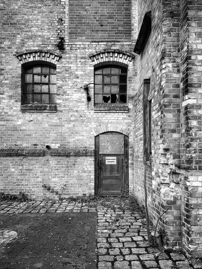 A b/w photograph of a building corner of the former Bremer Wollkämmerei (BWK). Above a locked wooden door with the sign "Do not enter! Danger to life!“ are two mullioned windows,
some of the panes are broken.