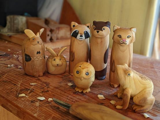 Groupe de sculptures en bois sur un plan de travail. Un monstre lapin, un lapin dodu, un raton laveur, une loutre, un chat et une grenouille. Devant eux, une petite chouette. Sur le plan de travail abimé il ya un couteau de sculpture et des copeaux. En arrière plan, flou, du bordel.