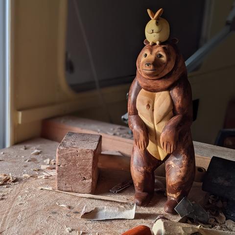 Sculpture d'un ours avec un lapin dodu posé sur sa tête. La sculpture est posée sur un établi encombré, un maillet, des copeaux, de la sciure, du papier à poncer.