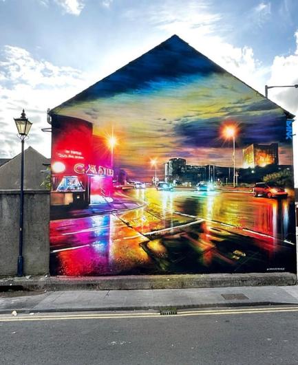 Streetartwall. On the outer wall of a one-story house was sprayed / painted the great mural of a city scene in the rain. The manga-style drawn scene of a Northern Irish city with neon signs, streets and evening sky looks very lively and is fantastically implemented. Some cars drive fast through the colorful streets and an ancient castle appears in the background. The lanterns shine, a cinema entrance can be seen and the scenery is reflected on the rain-soaked asphalt.