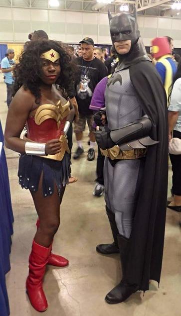 a dark skinned Black woman with long curly black hair dressed as Wonder Woman standing next to a tall white man dressed in a black and gray Batman costume at a comic convention