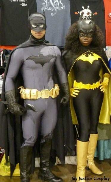 a Batman cosplayer dressed in a gray and black costume with a yellow belt, holding a batarang, next to a Batgirl cosplayer in a black and yellow costume, at a comic convention