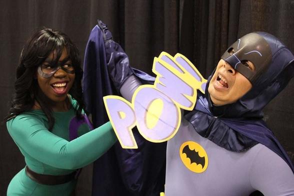 a smiling woman dressed in green with a purple question mark on her leotard (she is Query, an associate of the Riddler) punching Batman, who is wearing the gray and blue costume seen in the 1966 version. there is a 3 dimensional POW prop where her fist meets his face and he is falling over dramatically with his arms spread wide