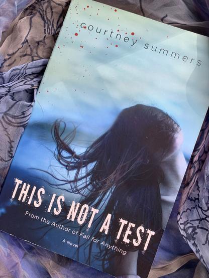 Pictured is This Is Not A Test (with cover art depicting a young woman, her face obscured by her hair) on top of a scarf in similar shades of blues and purples. Photo by AHS.