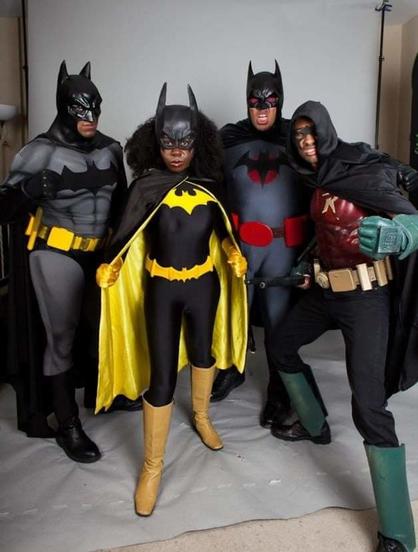 4 Black people dressed in cosplay. From left to right: Batman, in a black and gray costume with a yellow belt, Batgirl in a black and yellow costume, Batman (Flashpoint version) in a black, gray and red costume, and Robin, in a red and green costume with long pants and a long hooded black cape (cosplayer's own version)
