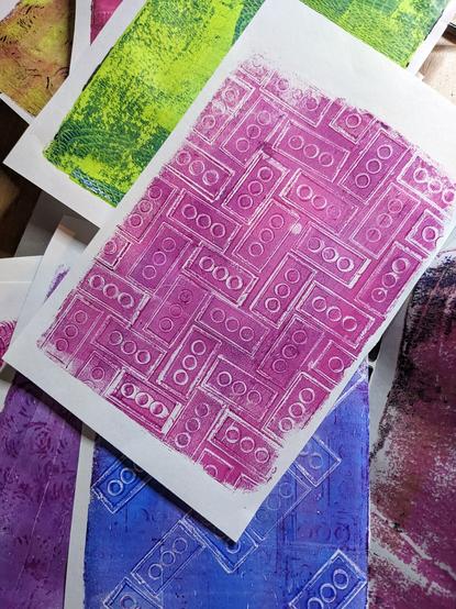 Stack of gel plate prints, the most prominent being a pink print of the underside of a Lego all over the page.