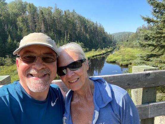 Two people wearing sunglasses are smiling. They are standing next to a railing. Behind that is a waterway surrounded by a deep green forest.