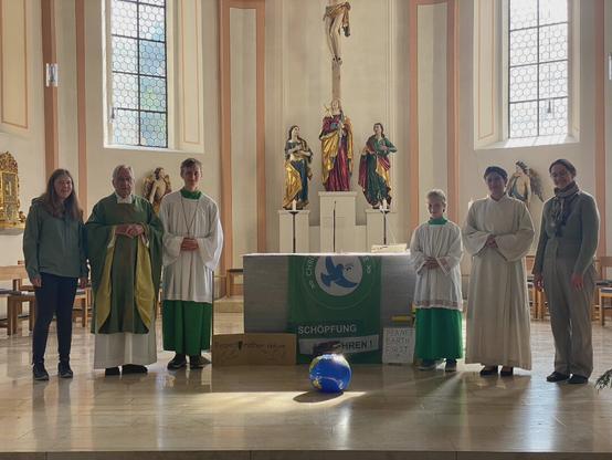 Sechs Personen, darunter ein Priester und drei weitere Personen in Messgewändern, stehen im Altarraum einer Kirche. Der Altar ist mit einer C4F-Fahne geschmückt, vor dem Altar liegt ein Ball, der aussieht wie eine Weltkugel.