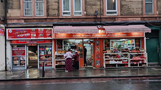 Bright orange front in Arabic, Asian, Fijian & African grocery store, with striped white & orange awning above it, shelves of fresh fruit and vegetables displayed outside, colours in contrast to rain & grey light