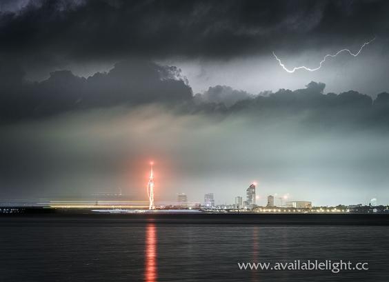 A night time view of the Portsmouth skyline from the other side of the Solent. The city is lit up and the most obvious building is the Spinnaker Tower. A red light shines brightly atop the Tower, a reflection from this is visible across the calm sea. The cloudy dark sky is illuminated by lightning, a small fork of which is visible in a clear patch.
