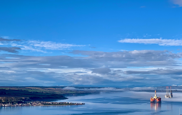 A blue sky layered with white and grey clouds, distant mountains, a haar across a still see. Two rigs centre with land on the left of the shot. A village of white houses strung out into the firth.