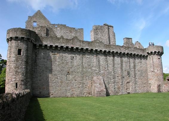 Craigmillar Castle. The image shows a grassy area in the foreground with a tall grey stone wall beyond it, with a circular tower at either end. The structure has battlements on top and there are gables and other parts of buildings projecting above the wall. Three people are standing on top of the right-hand tower. The scene is in sunlight.
