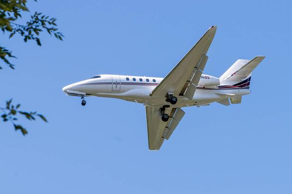 Image of a small jet in flight against a blue sky with a few leafed branches visible in the left frame. The jet is white with thin black and brown striping that extends from nose to tail. The jet's flaps and wheel are down indicating that it is on a landing approach.