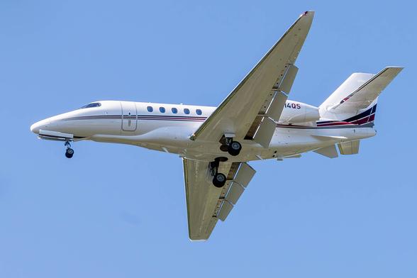 Image of a small jet in flight against a blue sky. The jet is white with thin black and brown striping that extends from nose to tail. The jet's flaps and wheel are down indicating that it is on a landing approach.