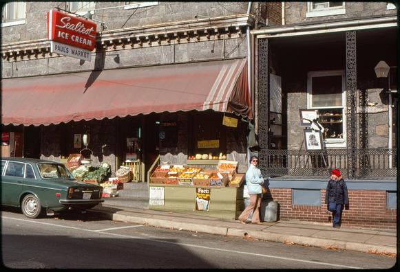 A small independent market fronts onto a narrow sidewalk. In front, bins of produce face the street. A faded red awning extends over the sidewalk. Above, a sign reads “Sealtest Ice Cream - Paul’s Market.” A hand-written sign in the window reads “Fresh Turkeys.” A green Volvo sedan is parked on the street. The porch of the building to the right is also visible, containing an easel holding artwork. Two people are on the sidewalk: a woman (looking toward the photographer) wearing a light blue coat, tan slacks, and a blue knit cap; and a boy in dark blue pants, a darker blue coat, and a red-and-blue knit cap.