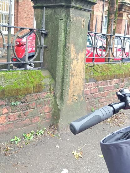An arrowhead OS benchmark cut into a stone (or possibly stone effect?) gatepost in a low red brick wall. Intricate railings fill the space above the wall.
The cut mark looks like it's been quickly engraved with a power tool. Not a pretty BM.
Photo taken from my bike and the handlebar is visible.