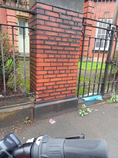 An arrowhead OS benchmark cut into the stone base of a red brick pillar in between sets of railings.
It's a nicely engraved BM which made me exclaim "Ooh, that's a nice one".
There is litter on the pavement.
My bike's righthand handlebar grip is visible as I took the photo from my bike.
