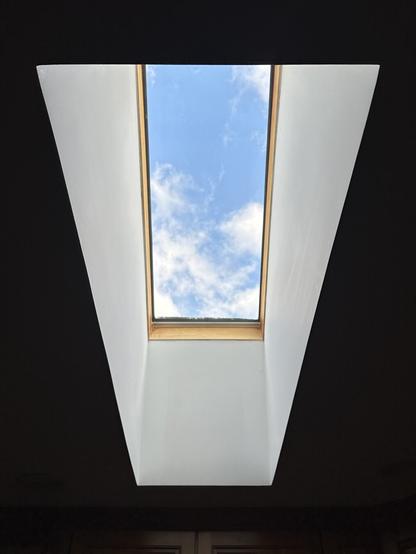 Looking upward through a long, narrow skylight, revealing a bright blue sky dotted with small white clouds.