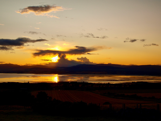 sunset over the Cromarty Firth. Orange and yellow hues. Sun behind dark clouds reflected in the water. A really dark shoreline foreground