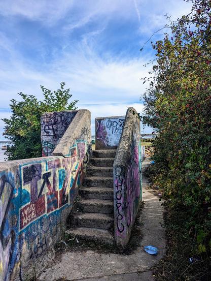 Below a blue sky with white fluffy clouds there's a stone path, a concrete wall rises with steps showing a curve and gap through to the left. There's a lower path to the right, and bushes on both sides.
