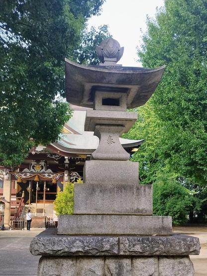 A big stone lantern on a massive pedestal in the foreground. In the background there is a reveler in front of the wooden shrine that is slightly obscured by trees.