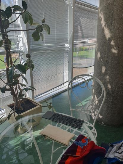 ein weißer stuhl mit verschnörkelter lehne vor einem glastisch, auf dem ein paar schreibutensilien liegen: mäppchen, bunte stoffhülle mit schreibttagebuch und klappbare tastatur, die mit dem handy zusammen gerade mein schreibutensil ist.