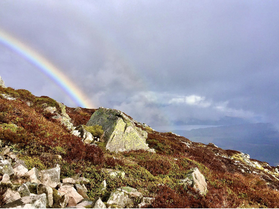 A moody grey sky with water centre right. A rainbow far left top corner with a hint of repeat. Steep side of a mountain with rocks and russet autumn broken in the foreground.