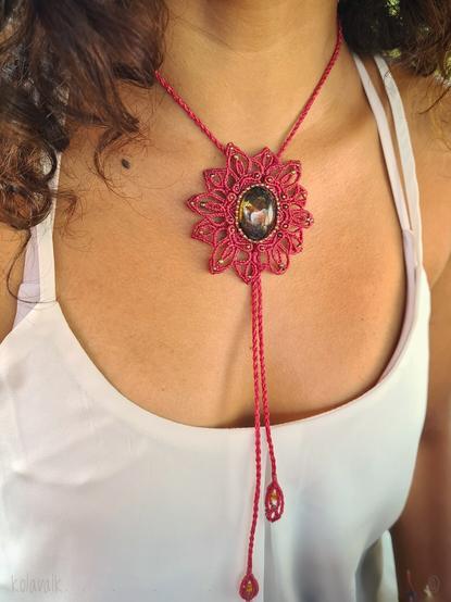 Photo couleur du cabochon d'ambre, couleur brun-orangé avec des inclusions +ou- opaques au milieu de la matière transparente, serti comme une "étoile-fleur" avec du fil rose fuchsia et quelques perles de métal de bronze, porté à même sa peau bronzée. On voit une partie de ses cheveux bruns bouclés et son débardeur blanc à fines bretelles.