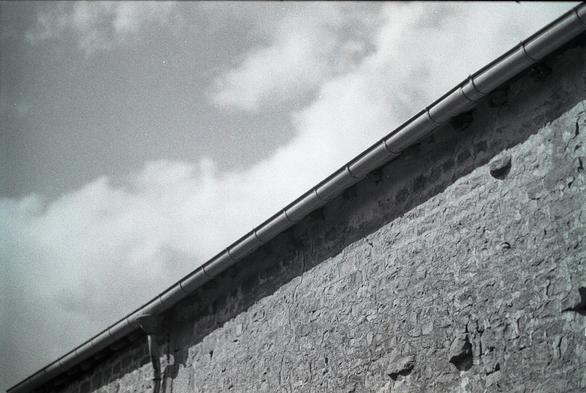 Black and white image divided from the lower left corner to the top right by a long gutter. The lower half of the frame is the sandstone wall that holds it, above the guttter a cloudy sky.