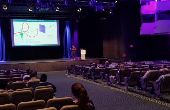 Max Frenzel on stage as a speaker in front of a plenary audience. He's poiting at a big projector slide, which shows a schematic of a helical THz pulse detected by a short probe pulse in a solid medium.