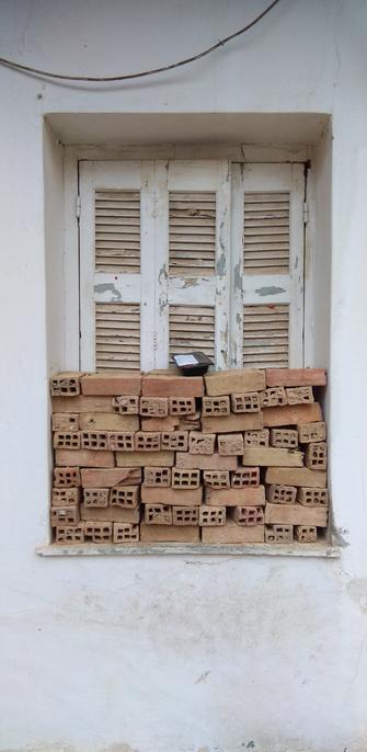 Window of empty old house, squatted quietly by homeless people during last winter. White wall, once white wooden shutters, closed. Half the window is blocked by bricks in rows. On top of them a food container, probably left there for the inhabitant(s). A cable forms a loose curb above the window.