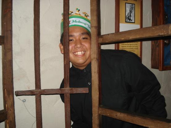 Me smiling and peering through a wooden cage. I'm wearing a black T-shirt and a green paper crown with the words "Medieval Times" on it.