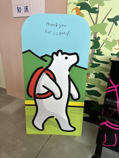 Tokosan wearing a backpack with some mountains in the background on a sign that says “thank you for coming!”
