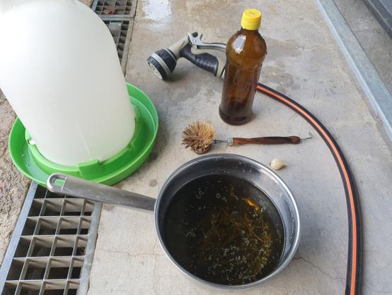 Auf dem Boden steht eine frisch gereinigte Hühnertränke, eine Flasche mit Apfelessig und ein Topf mit Wermuttee. Daneben liegt ein Wasserschlauch mit Brause, eine Spülbürste und eine Knoblauchzehe.