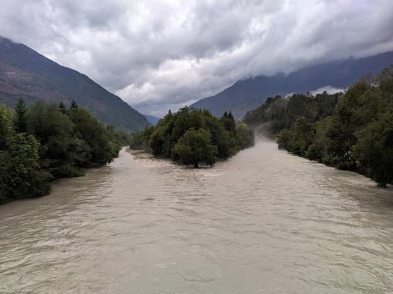 Ein Fluss teilt sich vor einer Insel, das Wasser braun, im Hintergrund Wolkendecke und Berge