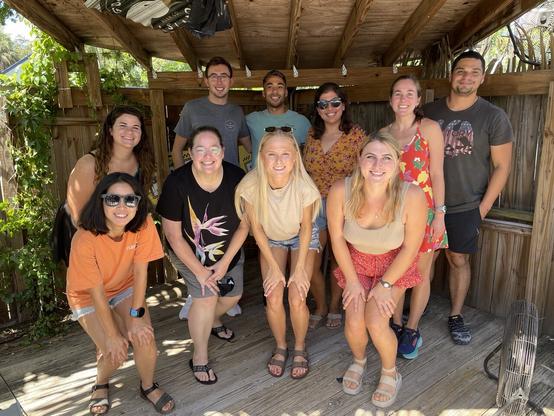 Group of ten people posing for the camera under a tiki hut on a hot, sunny day