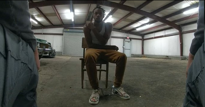 Photo of a black lad sat in a chair in the middle of an emopty warehouse infront of two white officers.