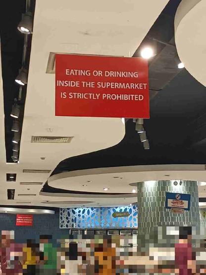 Photo shot inside a supermarket, showing a red sign hanging from the ceiling saying "Eating or drinking inside the supermarket is strictly prohibited."