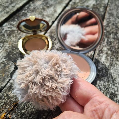 Photo de la houppette, ronde en laine duveteuse grise, glissée sur mes doigts grace à une petite anse intégrée. En arrière plan on voit deux poudriers ouverts dont l'un montre mes doigts en reflet dans son miroir intégré et l'autre a un petit coeur doré.