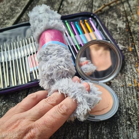 Photo de la houppette vue du dessus, ronde en laine duveteuse grise, glissée sur mes doigts grace à une petite anse intégrée. En arrière plan on voit un poudrier ouvert, un set de crochets multicolores et le reste de la pelote de laine synthétique grise duveteuse.