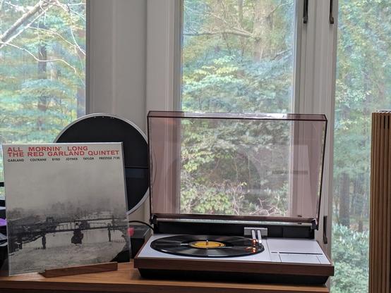 The Red Garland Quintet - All Mornin' Long LP cover.

A black and white picture of what looks to be the Hudson river in NYC, fog settling in, bridges over.

Title at the top in Red, participants in smaller type below in black.

The black LP plays on a vintage Bang & Olufsen Beogram 4000 linear-tracking turntable to the right. The dustcover is open, and the album, tonearm and surround are reflected in its underside.