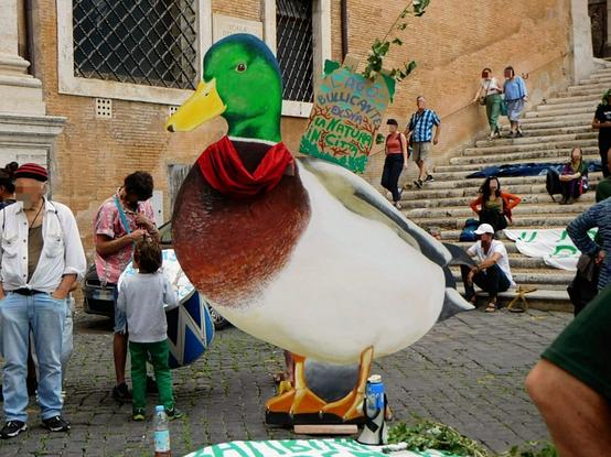 a big mallard duck hardback at a demonstration  (details on the rally in caption)    and some people next to it .   some are also  sitting  on the stairs behind
