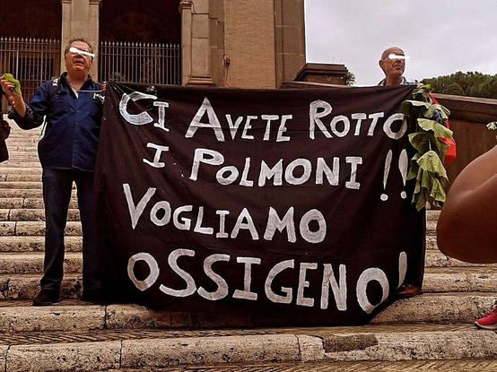 two men holding a big black banner  from the sides   at the rally ;      the white text is in italian :   " CI AVETE ROTTO I POLMONI !!   VOGLIAMO OSSIGENO! "   .      it's  a word play .    the literal meaning is:  " you broke our lungs!!  we want oxygen! " .     but  the first sentence also  sounds similar to   " ci avete rotto i coglioni ",   which would mean  'you're busting our balls'  (as in 'we had enough!')
