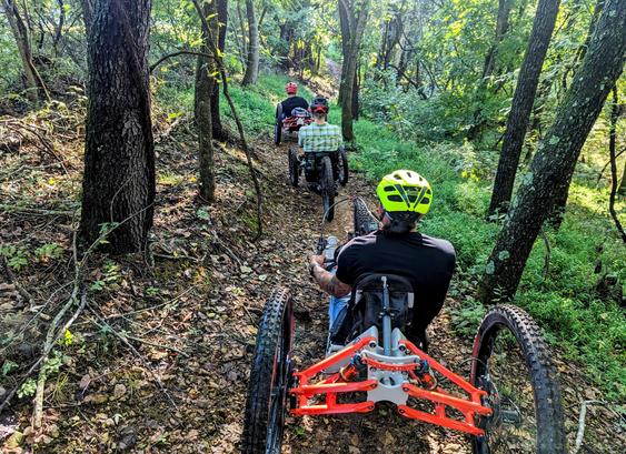 A train of adaptive mountain bike riders on a dirt trail through a forest