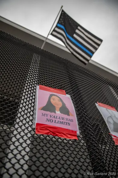 A photo of Jaahnavi Kandula taped to the front of the Seattle police union building. A "blue lives matter" looms atop the building. A large black chainlink fence keeps passers by at bay.