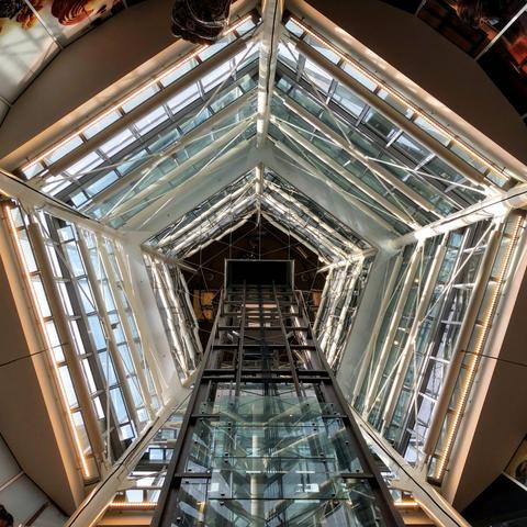 Upward view inside a glass and steel tower at a shopping mall in Amsterdam. It is pentagon shaped with a dark elevator mechanism seen at the bottom of the frame.