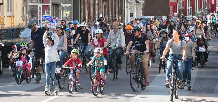 Foto der Kidical Mass Saarbrücken. Man sieht eine Straße, auf der viele Menschen samt Kinder auf Fahrrädern, Lastenrädern und ähnlichem zusammen unterwegs sind und offensichtluch Spaß haben. Das Wetter ist sonnig. Die Häuser spenden aber Schatten.